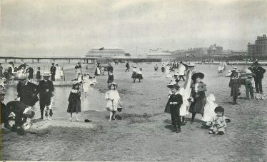 Black and white postcard showing children playing on a sandy beach with the pier in the background | Tuck Postcards database