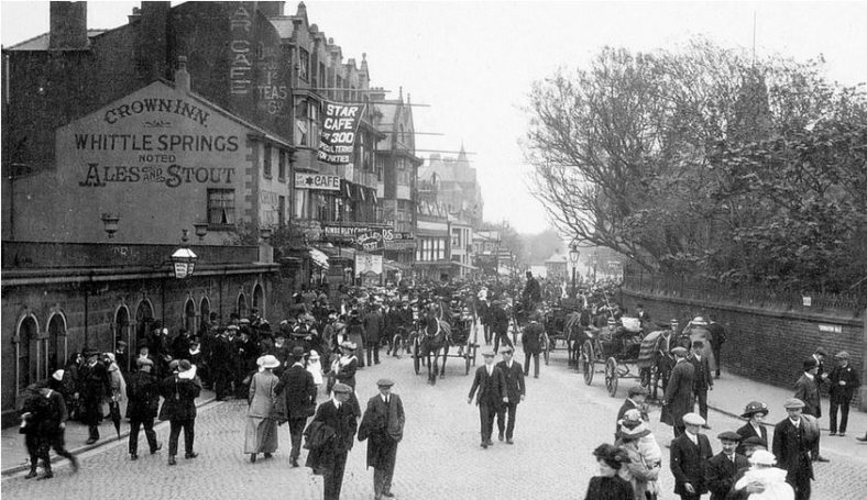Black and white photograph looking down Coronation Walk with lots of pedestrians, buildings on the left and a wall and trees on the right