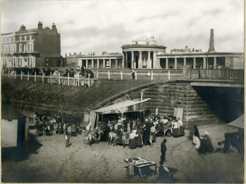 This early photo from the 1860s shows where Nevill Street tunnelled through the sand dunes under the Promenade and onto the beach. A long row of visitors - or residents - take in the sea air in front of the first seawater swimming baths. Below them is the hustle and bustle of stalls positioned to catch the tourists.  | The Atkinson, Southport